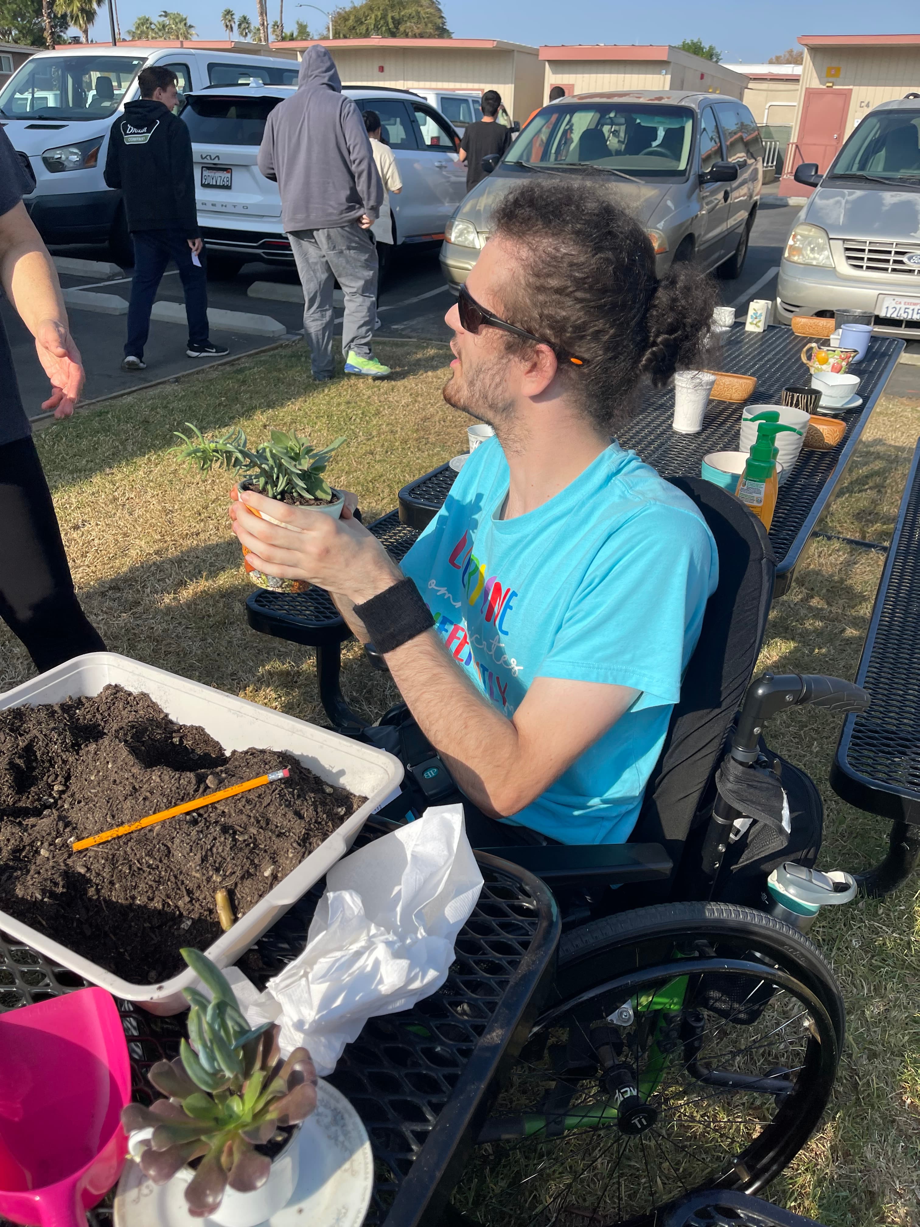 Man in a wheelchair holds a small succulent plant at an outdoor gardening table.