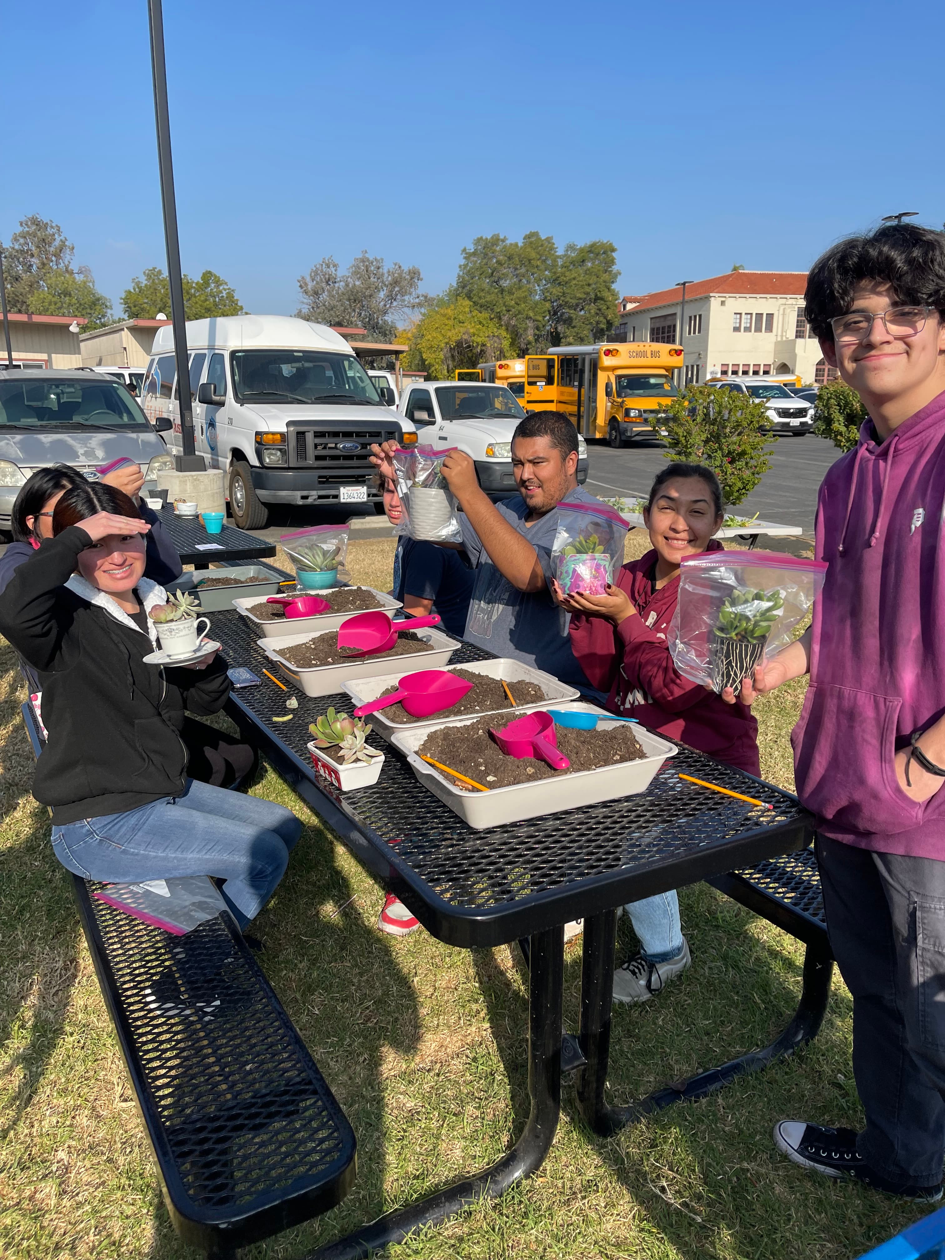Students smile while potting small succulent plants at a picnic table in a parking lot.