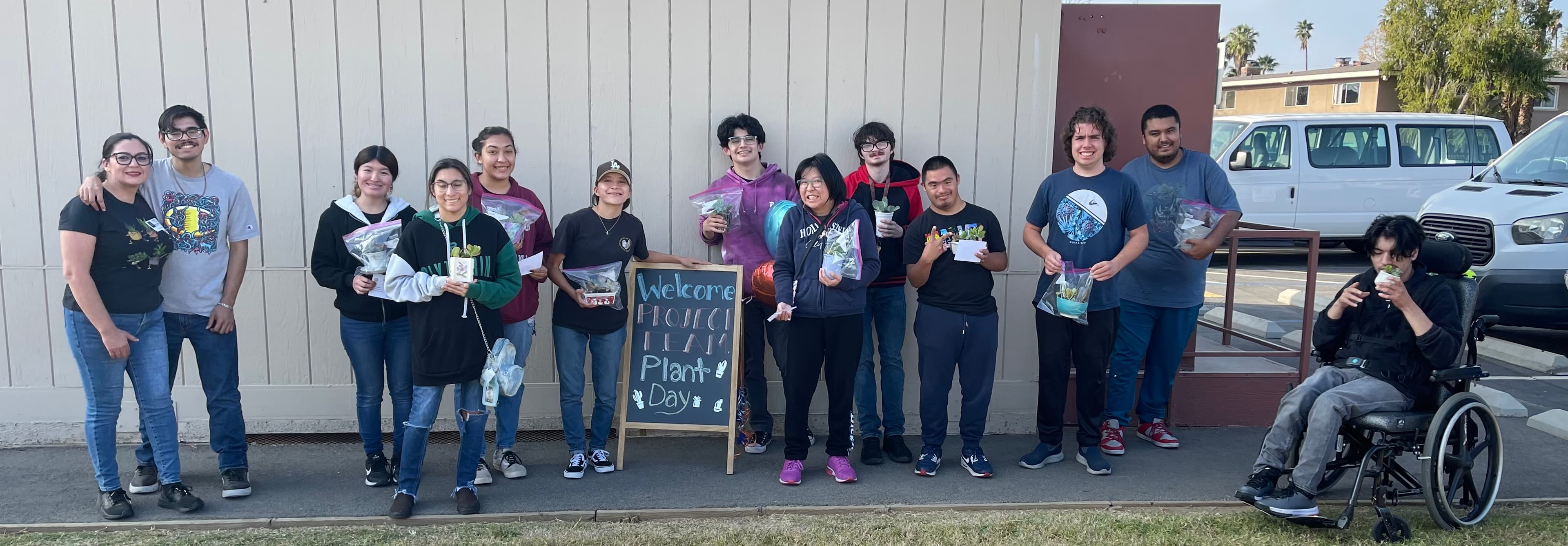 Diverse students holding small plants next to a Project Team Plant Day chalkboard sign.