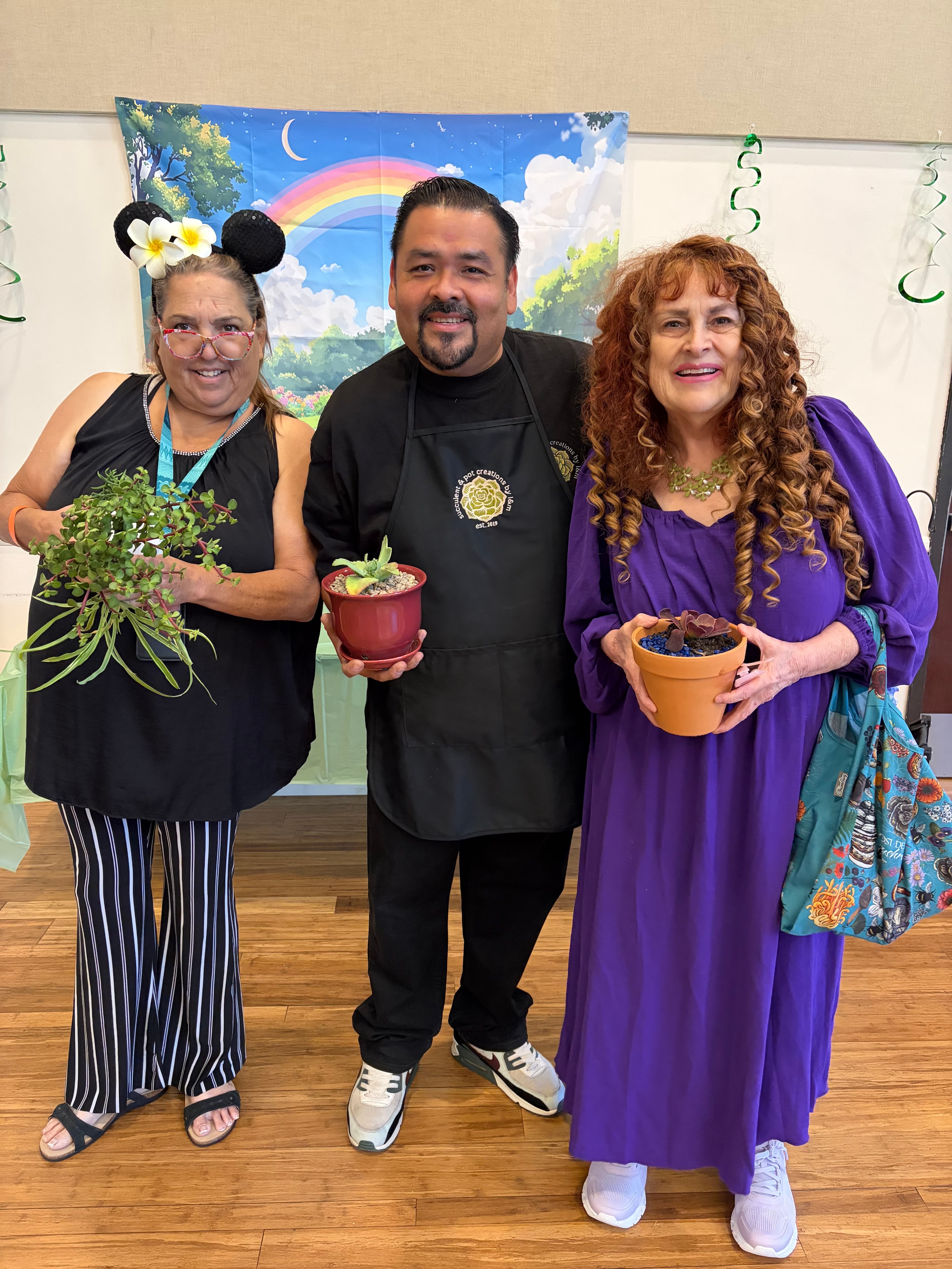 Three smiling people holding potted plants in front of a colorful rainbow and moon backdrop.