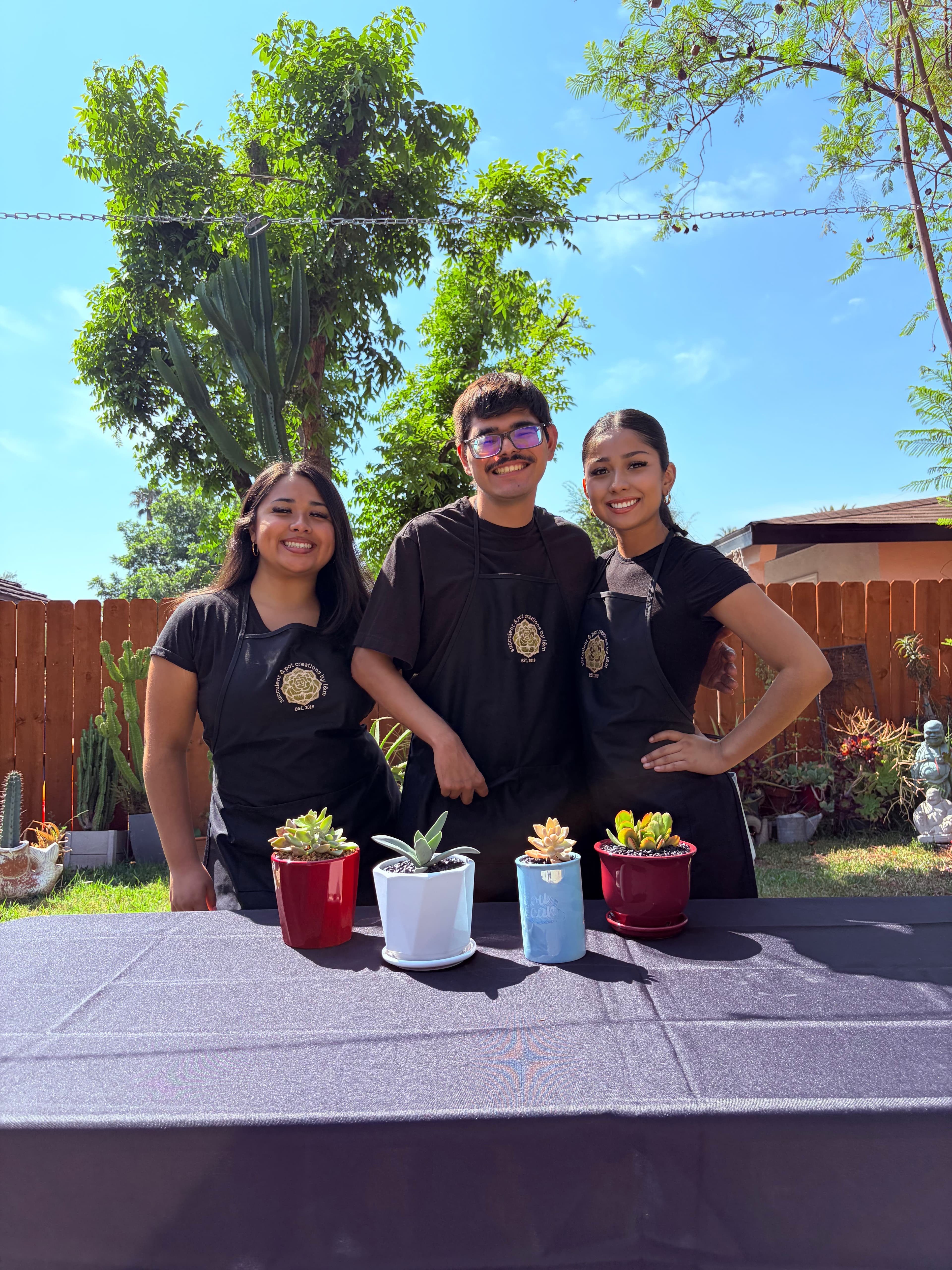 Three smiling people in black aprons stand behind a table displaying four potted succulents.