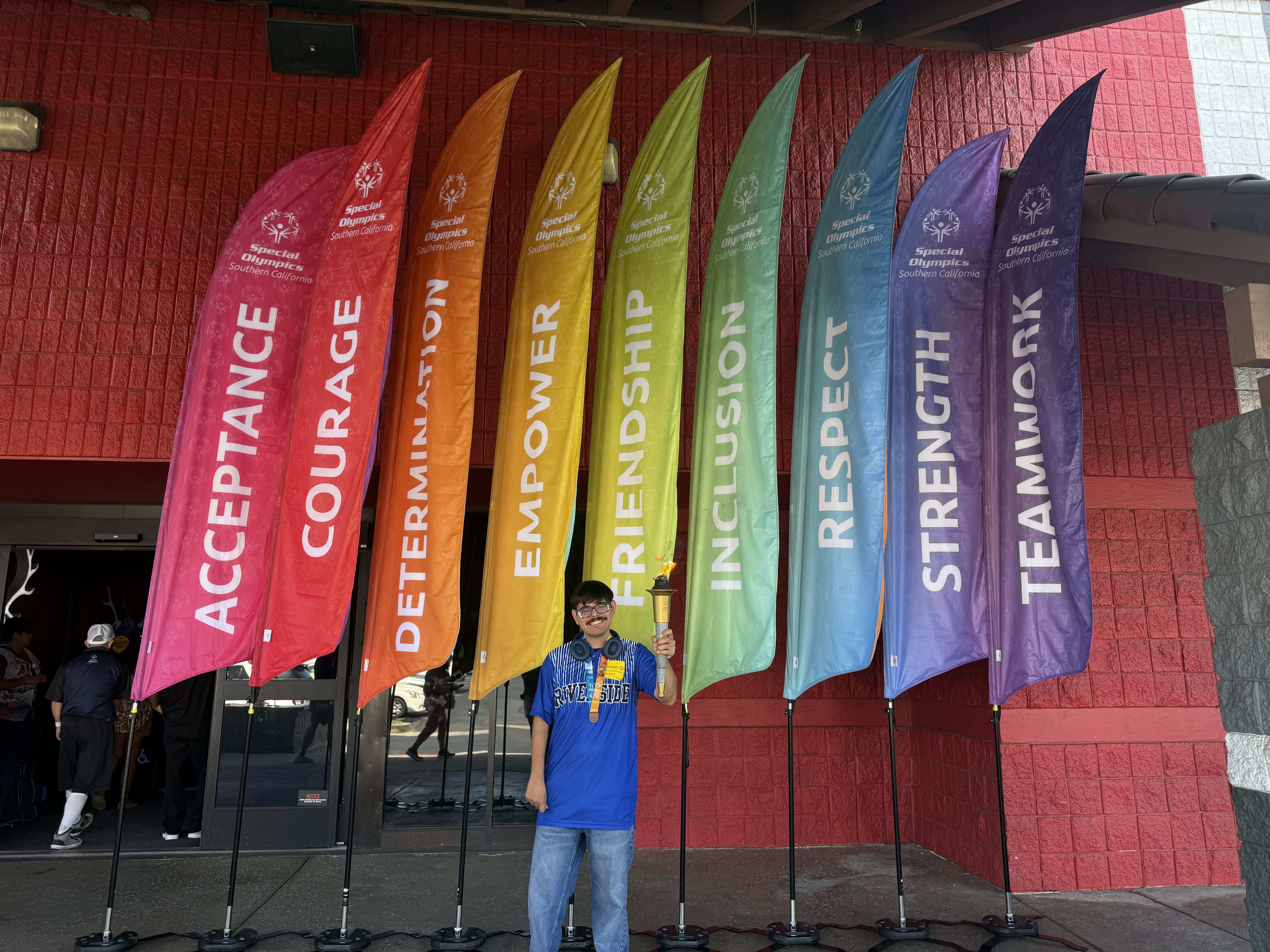 A man holding a torch stands before colorful Special Olympics flags displaying inspirational core values.